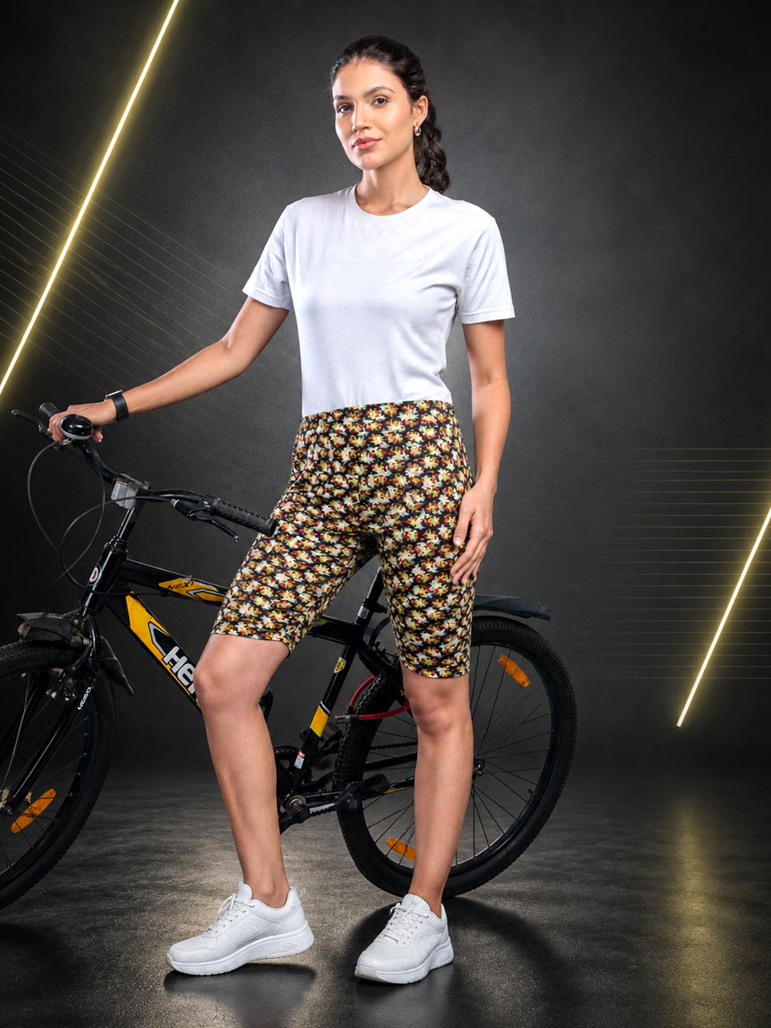 A woman stands next to a black bicycle in a studio setting, wearing a white t-shirt, high-waisted black shorts with a yellow and white floral pattern, and white sneakers.